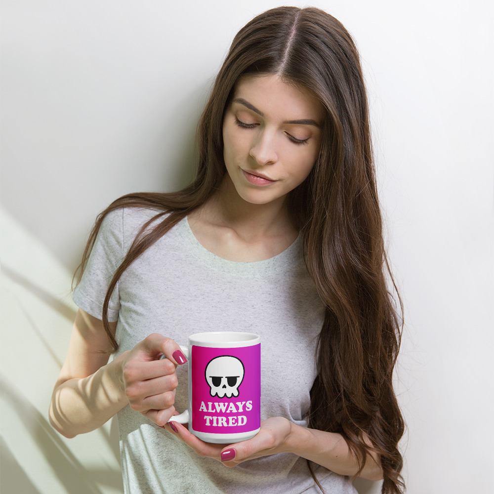 a person with long hair holds a tall white mug with a pink gradient background and it says always tired with a grumpy skull