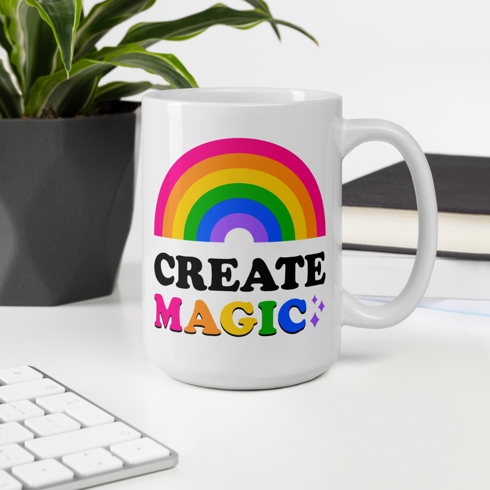 tall white mug that says create magic with a rainbow and colorful sparkles sitting on a desk with a keyboard, book, and plant
